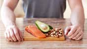 Junger Mann sitzt am Tisch, auf dem ein Brett mit Lachs, Avocado und Nüssen liegt.