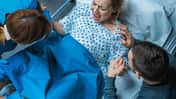 Draufsicht: Man sieht eine schreiende Gebärende in einem Klinikbett, neben ihr steht ihr Mann