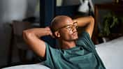 A young man sits in his lounge on the couch while relaxing with his hands behind his head. Stock photo