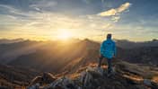 Man on stone on the hill and beautiful mountains in haze at colorful sunset in autumn. Dolomites, Italy. Sporty guy, mountain ridges in fog, orange grass and trees, blue sky with sun in fall. Hiking