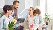 Eine Frau sitzt auf dem Sofa, ihr Mann und ihre Kinder reichen ihr Blumen und Schokolade und eine Karte zum Muttertag