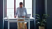 Ernsthafter Geschäftsmann, der mit Laptop und einer Tasse Kaffee am Schreibtisch steht und Notizen in ein Notizbuch schreibt.
