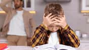 Ein kleiner Junge sitzt angestrengt über einem Schulbuch, im Hintergrund sieht man einen verzweifelten Vater
