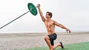 Side view of a young muscular man training on the beach shirtless doing landmine overhead press with barbell in kneeling position.