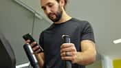 Ein Mann trainiert auf einem Crosstrainer im Fitnessstudio, hält einen Griff des Geräts und blickt auf sein Smartphone. Er trägt einen Fitnessring