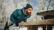 Mann trainiert draußen und macht Liegestütze an einer Holzbank im verschneiten Park. Mann trainiert draußen und macht Liegestütze an einer Holzbank im verschneiten Park.