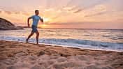 Ein sportlicher Mann läuft am Strand während im Hintergrund die Sonne untergeht Ein sportlicher Mann läuft am Strand während im Hintergrund die Sonne untergeht