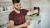 Junger Mann steht in der Küche und schenkt sich einen frisch gemixten Proteinshake in ein Glas ein