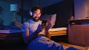 Mann mit Kopfhörern sitzt im dunklen Schlafzimmer an sein Bett gelehnt und schaut lachend auf sein Smartphone