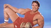 Arnold Schwarzenegger posiert vor einem blauen Hintergrund in einem roten Sportoutfit mit einer Kurzhantel