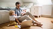 Ein Vater sitzt vor einem Sofa, mit einem aufgeklappten Notebook und zwei kleinen Kindern an seiner Seite